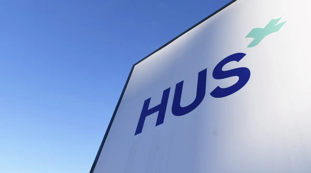 Kuvassa näkyy vaaleansinistä taivasta ja seinä, jossa on logo HUS.