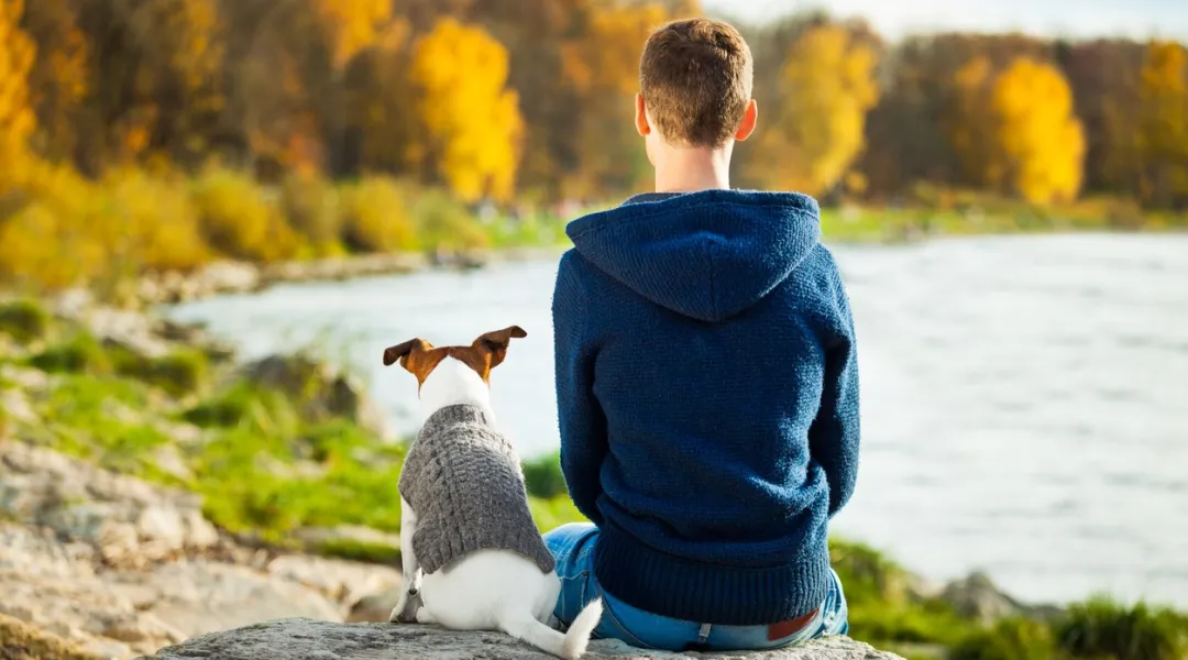 mies kuvattuna tapaapäin hänen istuessaan  veden äärellä koiran kanssa syksyllä