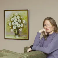 Nainen katsoo hymyillen kameraan, toinen käsi poskella. Vieressä seinällä on kukkataulu.