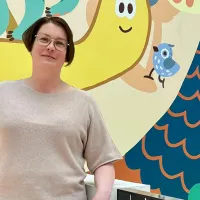Leena Lauri seisoo kuvassa ja katsoo kameraan. Tausta on piirretty ja siinä on iloisia värejä. 