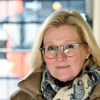 Silmälasipäinen nainen katsoo hymyillen, hänen yllään on talvitakki.