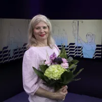 Nainen seisoo ja hymyilee. Hänellä on kädessään kukkakimppu.