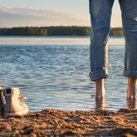 Kuvassa näkyy ihmisen jalat takaapäin seisomassa rannalla ilman kenkiä, jotka ovat hänen jalkojensa vieressä. Maisema on kauniin kesäinen. 