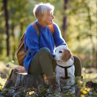Vanhempi nainen istuu koiran kanssa metsässä.