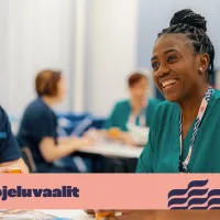 Vaaleatukkainen nainen ja tummaihoinen nainen istuvat ja hymyilevät. Kuvassa on teksti &quot;työsuojeluvaalit&quot;.