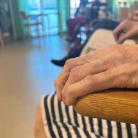 Vanhuksen käsi lepää tuolin selkänojalla. Vanhukset istuvat ringissä palvelutalon yhteisessä tilassa.