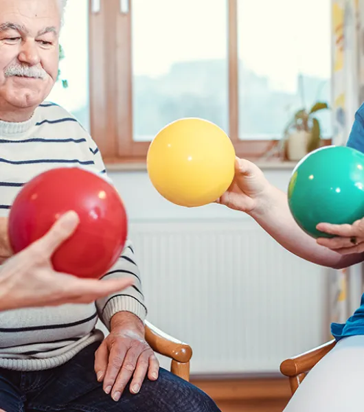 Kaksi iäkästä henkilöä ja hoitajaa pelaavat yhdessä erivärisillä palloilla.