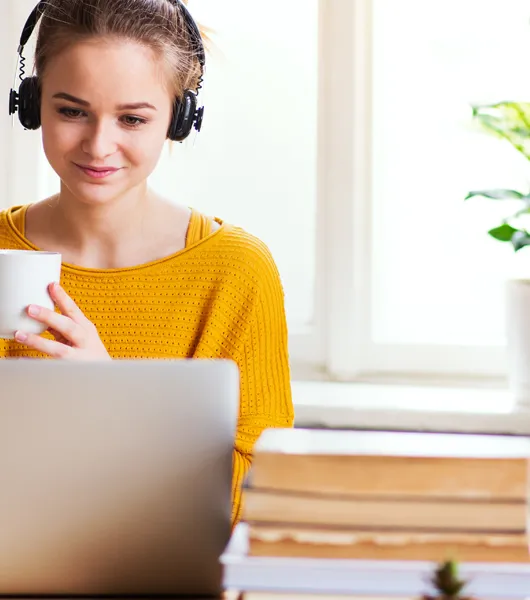 Nuori nainen istuu tietokoneen ääressä ja pitelee käsissään kahvikuppia.