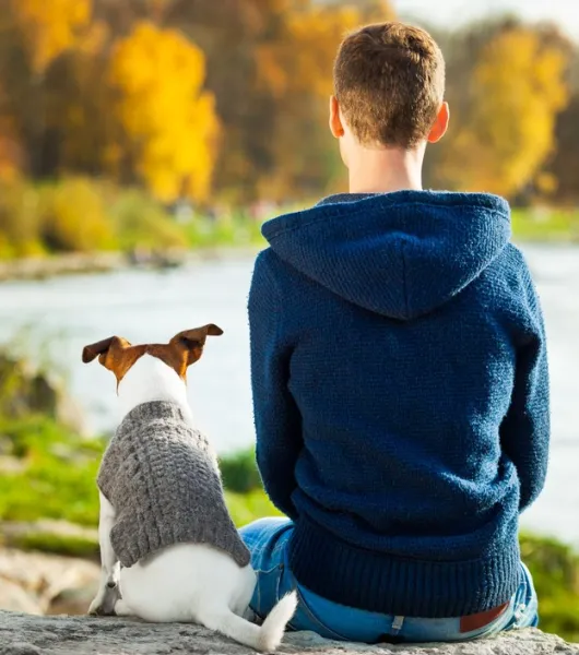 mies kuvattuna tapaapäin hänen istuessaan  veden äärellä koiran kanssa syksyllä