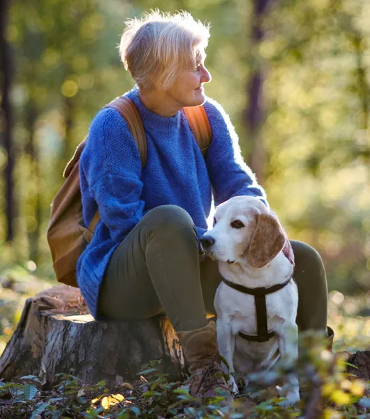 Vanhempi nainen istuu koiran kanssa metsässä.