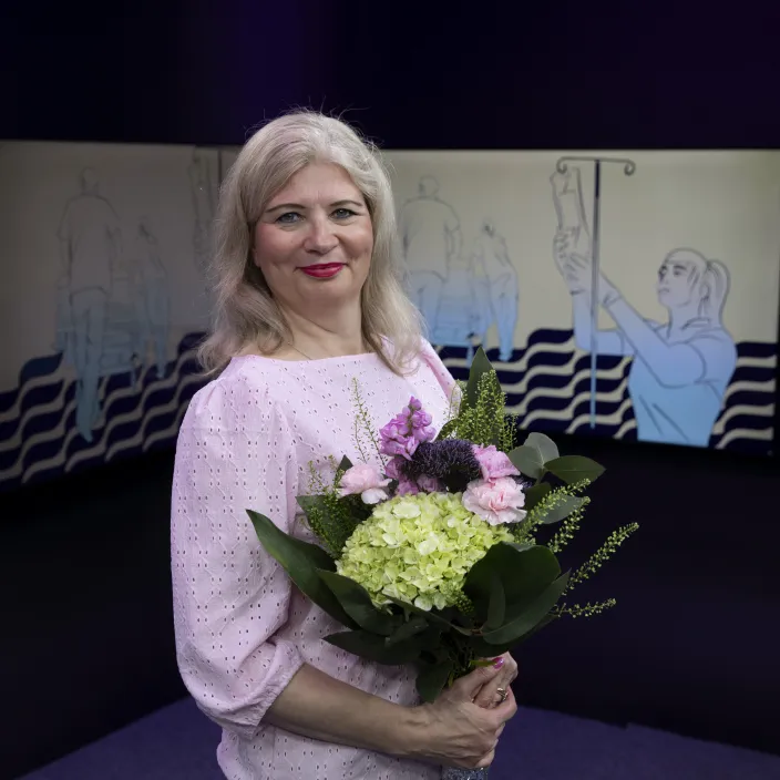 Nainen seisoo ja hymyilee. Hänellä on kädessään kukkakimppu.