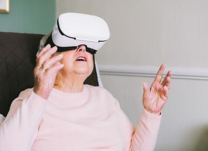 Vanhempi nainen istuu tuolissa vr-lasit päässään ja kurottelee käsillä ylöspäin.