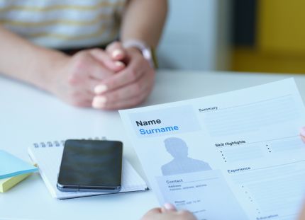 Henkilö istuu työhaastattelussa. Etualalla toinen henkilö pitää käsissään ansioluetteloa, jossa lukee &quot;Name Surname&quot;. Pöydällä on muistilehtiö, älypuhelin ja värikkäitä muistilappuja.