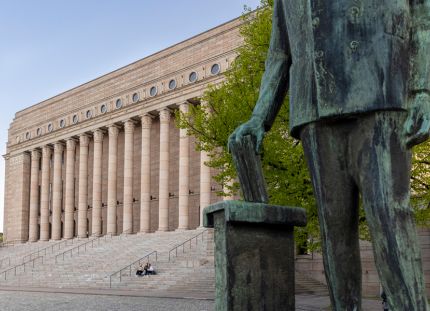 Eduskuntatalo Helsingissä kevätauringossa. Etualalla näkyy työväenjohtaja Yrjö Mäkelinin patsas, jonka hahmo pudottaa paperin vaaliuurnaan.