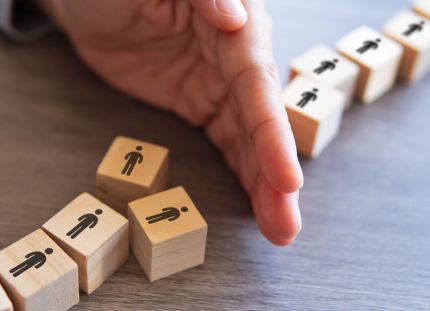 Laboratorioyhtiö Fimlab on aloittanut muutosneuvottelut, jotka voivat johtaa 30 henkilötyövuoden vähentämiseen. Käsi estää puisista palikoista muodostetun ihmisjonon etenemistä, yksi palikka on kaatunut muiden edessä.