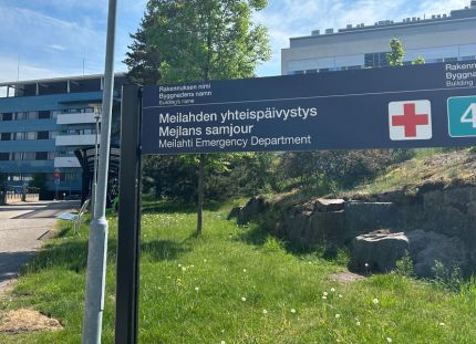 Meilahden sairaala-alue kesällä kuvattuna. Päivystys-kyltti nurmikolla. Hyvinvointialueiden säästöt ja rekrytointikiellot kiristävät sote-alan kesää. 