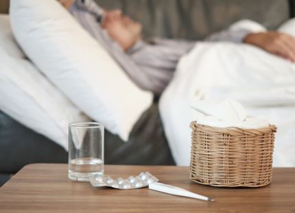 Influenssa jyllää, mutta rokote suojaa vakavalta taudilta. Sairastunut mies makaa sohvalla, pöydällä vesilasi, särkylääkkeitä ja kuumemittari.