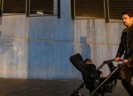Isä työntämässä rattaissa pientä lasta. Taustalla betoninen seinä.