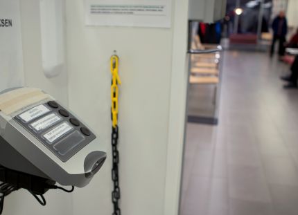 Odotustilassa vuoronumerolaite ja potilaita käytävän reunalla istumassa.