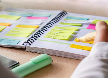 Henkilö kirjoittaa keltaisella korostuskynällä muistivihkoon, joka on täytetty värikkäillä post-it-lapuilla ja tehtävälistoilla. Vihko on avoinna pöydällä, jonka ääressä näkyy myös toinen korostuskynä ja älypuhelin.