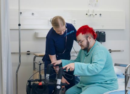 Hoitaja auttamassa potilasta nousemaan kävelytuen avulla ylös sairaalasängystä.