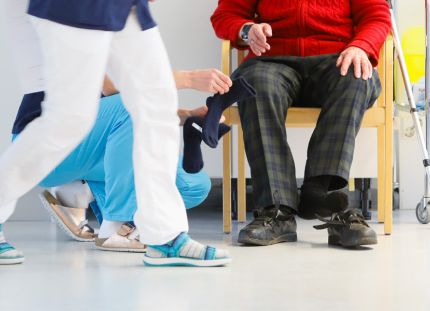 Kiireisiä hoitajia sairaalan käytävällä. Vanhus istuu tuolissa.