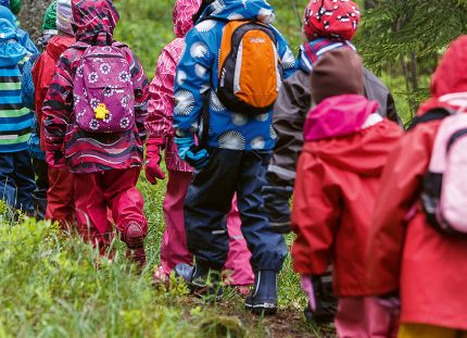 Päiväkotilapset kävelevät metsässä jonossa sadevarusteissa.