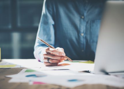 Henkilö laatimassa tietokoneella suunnitelmaa ja hahmottelee samalla kynällä paperille.