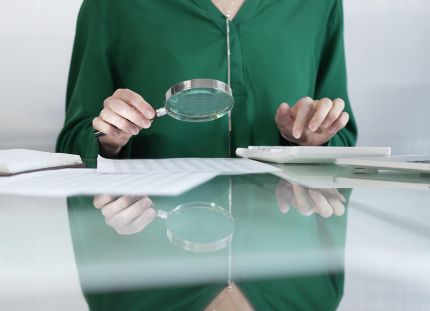 Ihminen istuu pöydän ääressä ja tutkii suurennuslasilla papereita. 