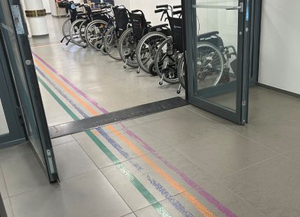Kuva sairaalan aulatilasta, jossa eri väriset viivat johdattavat kävijän oikeaan paikkaan. Taustalla seinustalla pyörätuoleja.