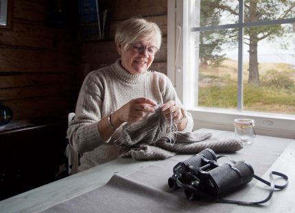 Tuovi Anttila neuloo mökillä pöydän ääressä ikkunan edessä.