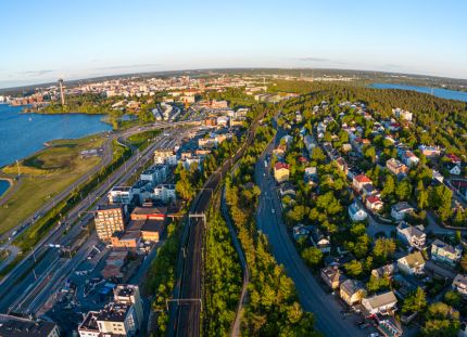 Ilmakuva Tampereen kaupungista kesäiltana, etualalla rautatie ja asuinalue värikkäine taloineen, vasemmalla suuri vesialue ja satama-alue, taustalla keskustan rakennuksia ja näkötorni, oikealla toinen vesialue ja vihreitä metsäalueita.