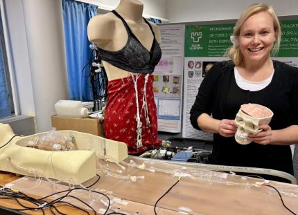 Yliopistotutkija Mariella Särestöniemi Human Body Twin -laboratoriossa, jossa kehitetään teknologiaa, joka mahdollistaa terveydentilan seurannan ja arvioinnin kannettavilla ja päälle puettavilla mittalaitteilla. Kädessään hänellä on aivomalli, jota käytetään signaalin etenemisen tutkimiseen aivokudoksessa, ja taustalla nukella on rintasyövän tunnistukseen kehitettävät monitorointiliivit. 