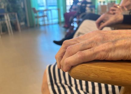 Vanhuksen käsi lepää tuolin selkänojalla. Vanhukset istuvat ringissä palvelutalon yhteisessä tilassa.