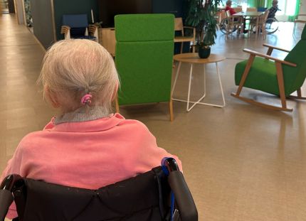 Vanhus istuu pyörätuolissa selin kameraan hoivakodin aulatilassa.