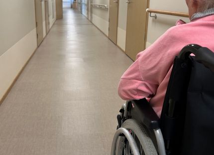 Vanhus sairaalan käytävällä pyörätuolissa. Puutteellinen kielitaito kuormittaa vanhusten hoidon työyhteisöjä ja vaarantaa potilasturvallisuutta.