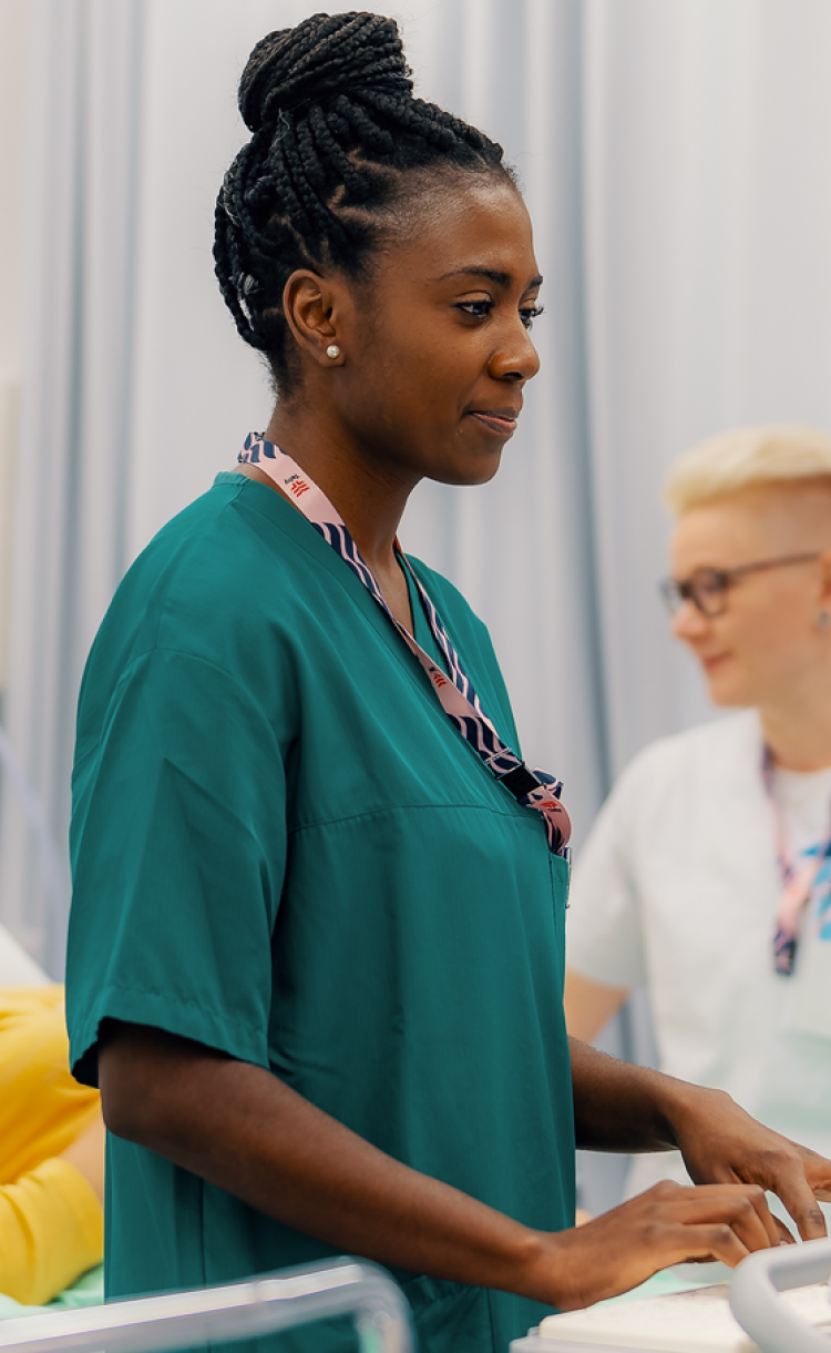 Kuvassa hoitaja käyttää monitoria ja taustalla on potilas ja toinen hoitaja