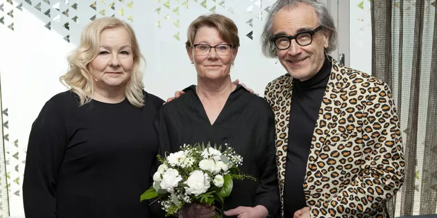 Kaksi naista ja mies hymyilevät. Keskellä olevalla naisella on kukkakimppu kädessään.