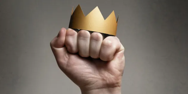 Kuvassa on käsi nyrkissä, jonka päällä on kultainen kruunu.