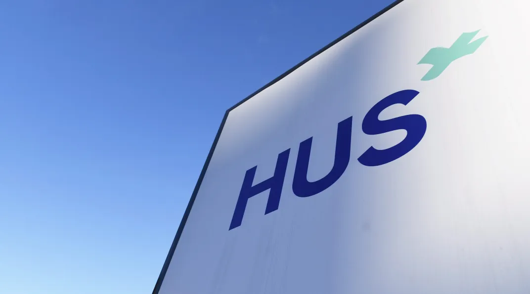 Kuvassa näkyy vaaleansinistä taivasta ja seinä, jossa on logo HUS.