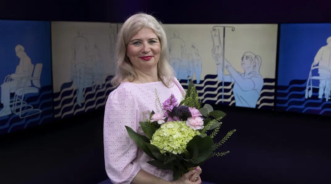 Nainen seisoo ja hymyilee. Hänellä on kädessään kukkakimppu.