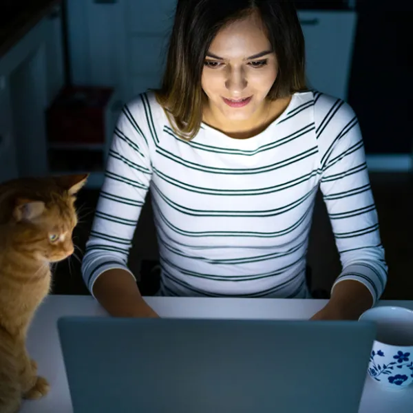Nainen käyttää tietokonetta illalla pimeässä keittiössä kissan kanssa